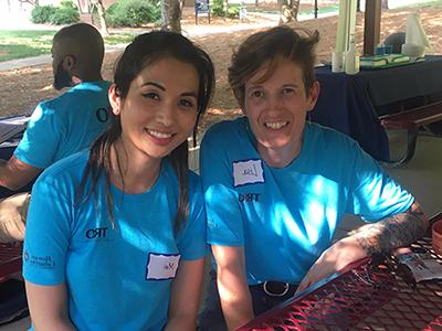 Two people in blue t-shirts sitting at an outdoor picnic table, smiling.